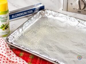 Large rimmed baking sheet lined with aluminum foil and coated with nonstick cooking spray, ready for roasting Brussels sprouts and apples. A can of olive oil cooking spray, aluminum foil box, and oven temperature dial set to 425°F are visible on a marble countertop with red patterned kitchen linens.