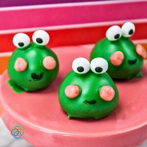 Frog Oreo Truffles for Halloween | Irish American Mom Three adorable green Frog Oreo Ball truffles decorated with large candy eyes, pink candy cheeks, and little black icing smiles, arranged on a pink serving plate against a colorful pink and red striped background.