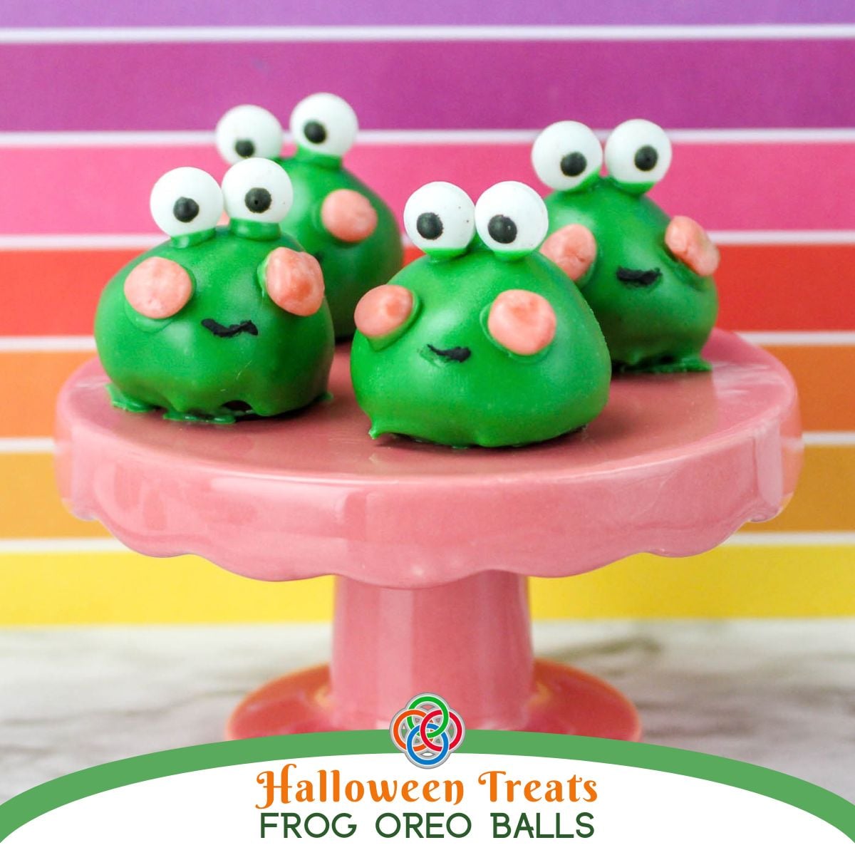 Three adorable green frog Oreo truffles with white candy eyes, pink candy cheeks, and black icing smiles displayed on a pink pedestal cake stand against a colorful pink, purple, and orange striped background over a text banner.