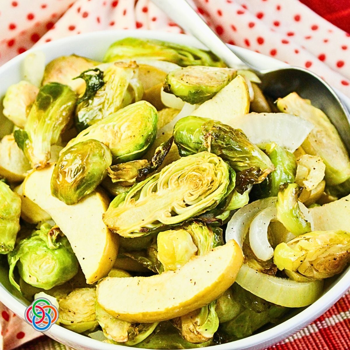 Close-up of a white serving bowl filled with roasted Brussels sprouts, sliced Granny Smith apples, and sweet onions. The sprouts have lightly browned edges, and a silver serving spoon rests inside the bowl against a red and white polka-dot napkin and red plaid tablecloth.