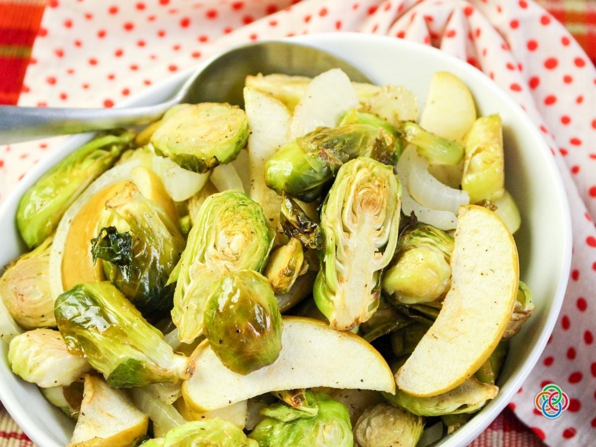 White bowl filled with roasted Brussels sprouts, sliced Granny Smith apples, and sweet onions, served with a silver spoon on a red plaid tablecloth and red polka-dot napkin.