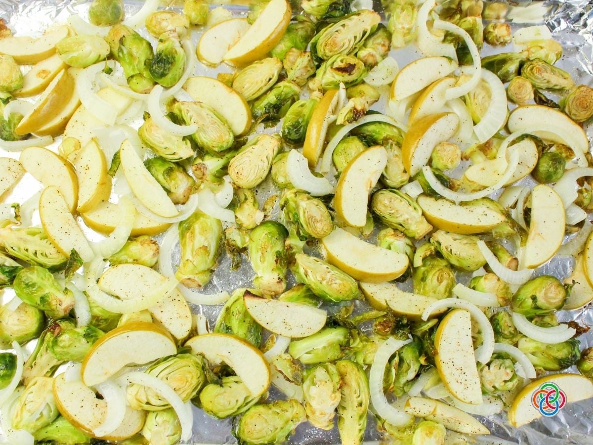 Overhead view of roasted Brussels sprouts, sliced apples, and sweet onion rings spread evenly across an aluminum foil-lined baking sheet. The halved Brussels sprouts have lightly browned edges, while the apple wedges and onions are tender and seasoned with black pepper.