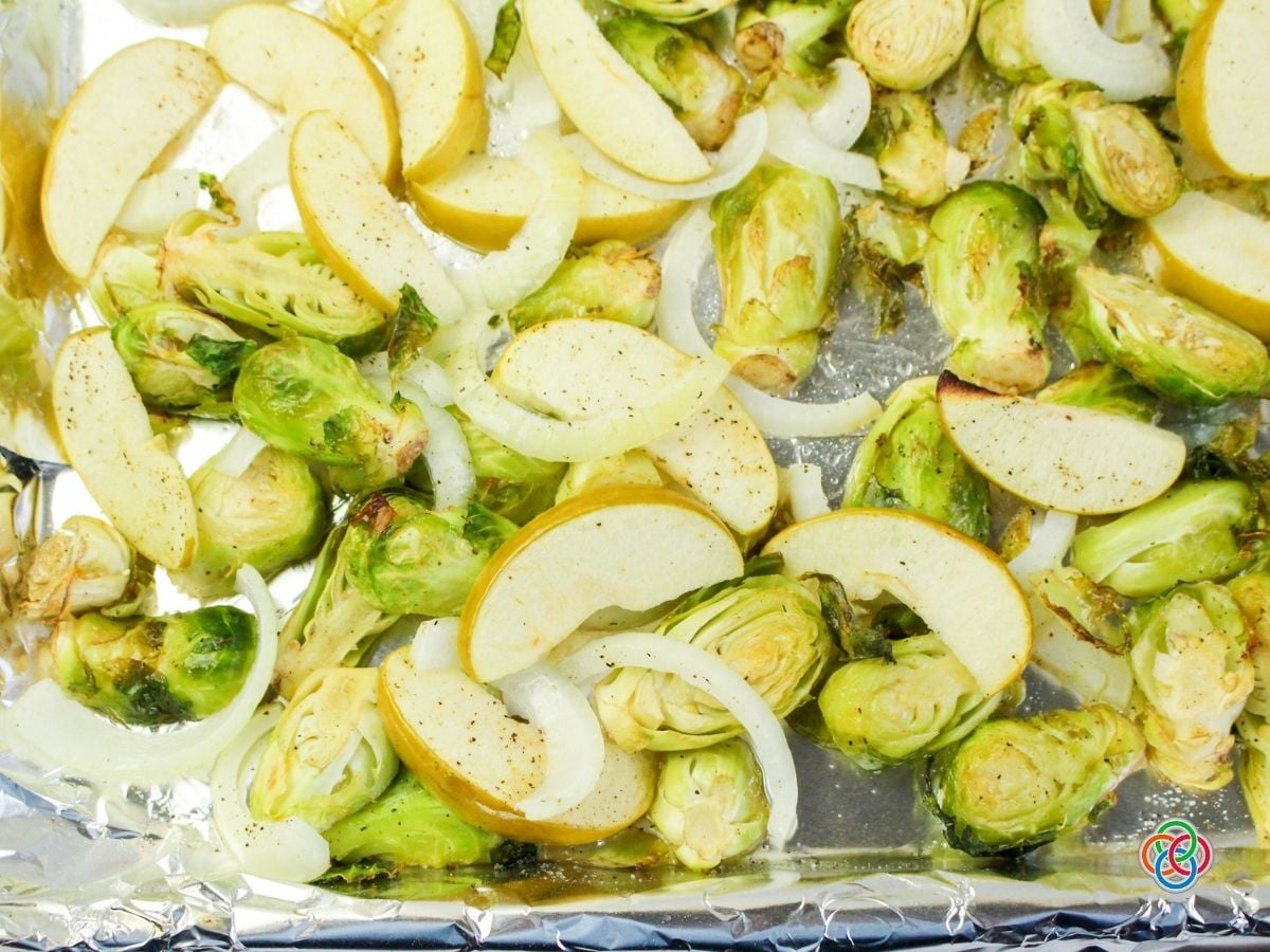 Close-up of roasted Brussels sprouts, sliced apples, and sweet onions seasoned with black pepper and spread across an aluminum foil-lined baking sheet. The Brussels sprouts have lightly browned edges, and the apple wedges are beginning to soften.