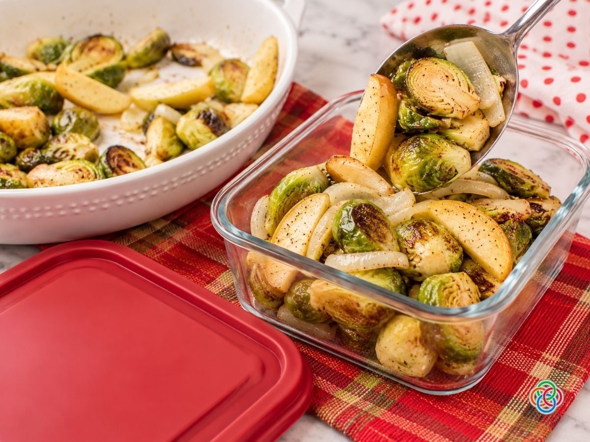 Roasted Brussels sprouts, golden apple wedges, and sweet onions being spooned from a white serving dish into a rectangular glass food storage container. A matching red lid rests beside the container on a festive red plaid tablecloth, with a red polka-dot napkin in the background.