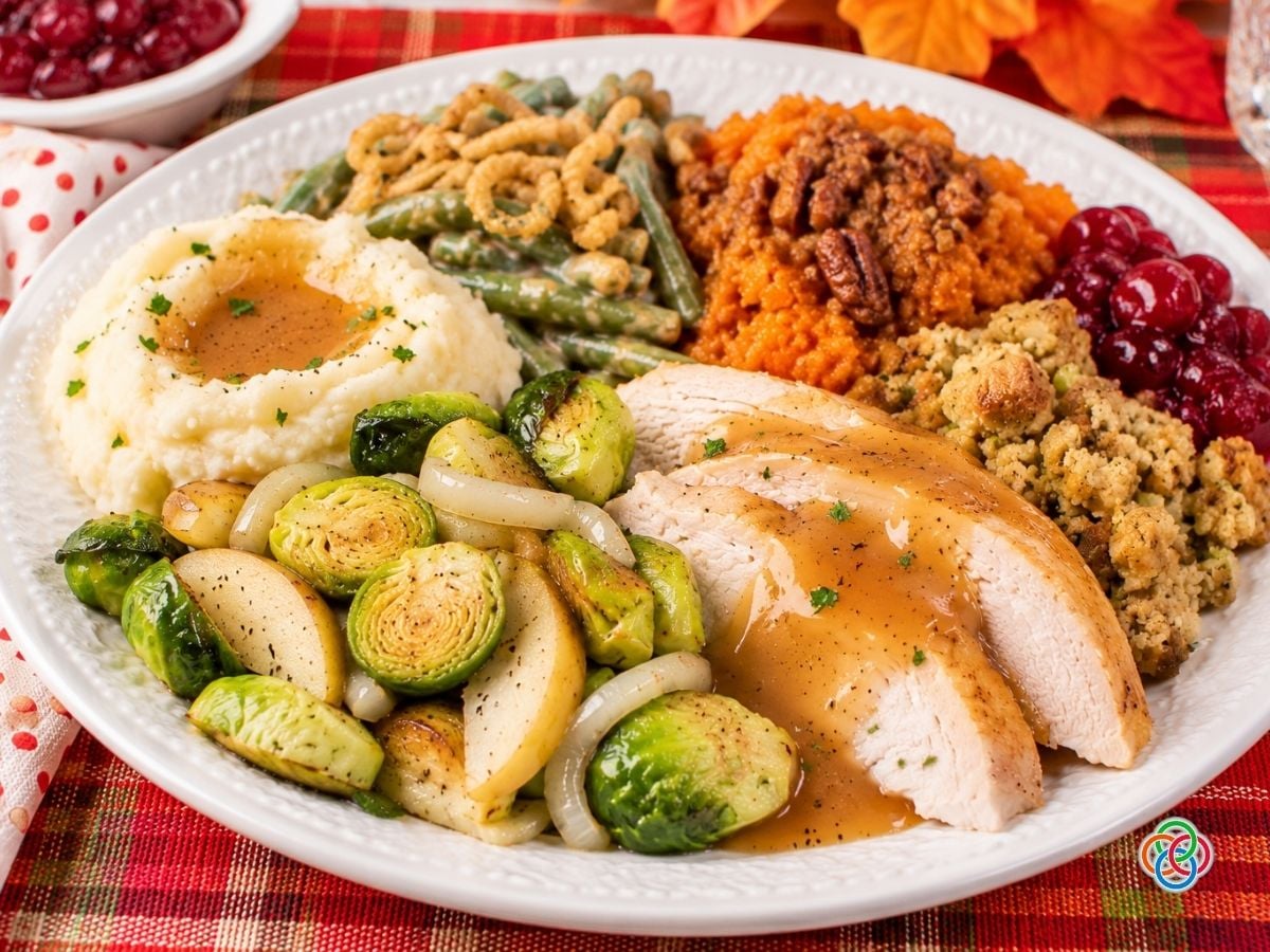 Traditional Thanksgiving dinner served on a white plate featuring sliced roast turkey with gravy, roasted Brussels sprouts with apples and onions, creamy mashed potatoes, herb stuffing, green bean casserole, pecan-topped sweet potato casserole, and cranberry sauce on a festive red plaid tablecloth.