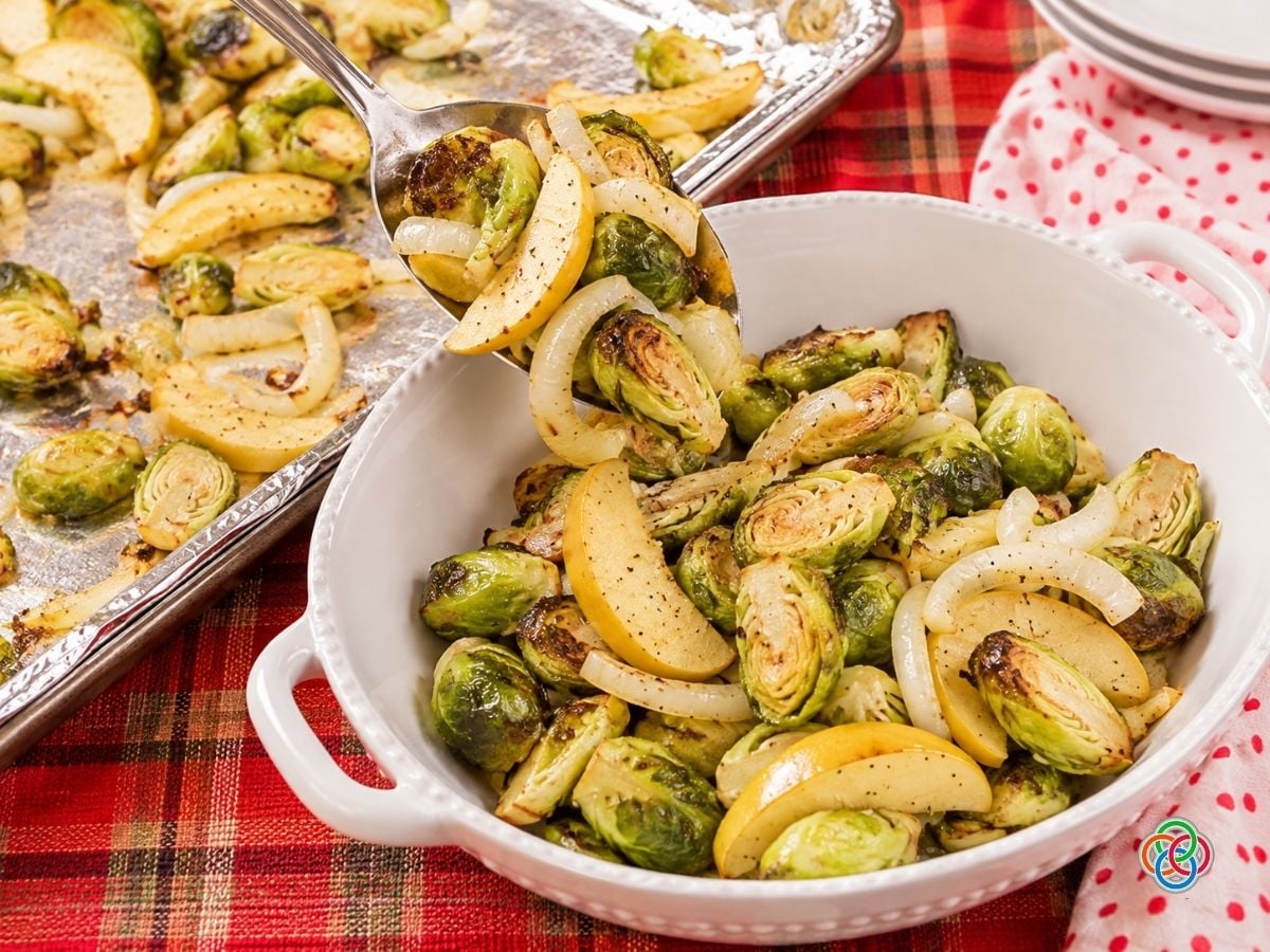 Roasted Brussels sprouts, golden apple wedges, and sweet onion slices being spooned from a foil-lined baking tray into a large white ceramic serving dish. The vegetables have lightly caramelized edges and are arranged on a festive red plaid tablecloth beside a white napkin with red polka dots.