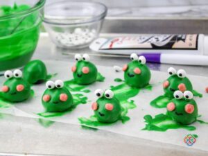 Using black icing to create frog mouth | Irish American Mom Green edible frogs on a baking sheet with candy eyes, pink chkkes and icing moutns