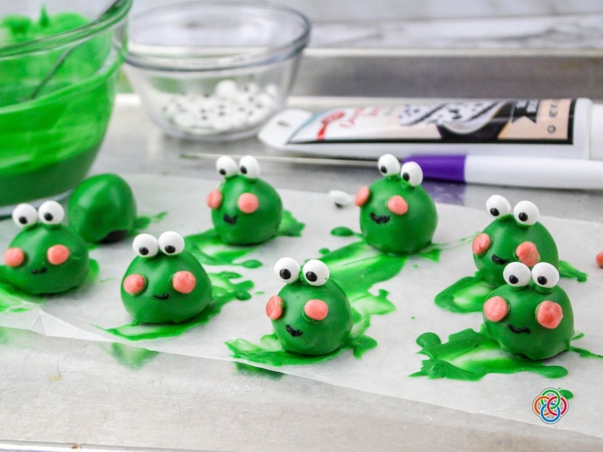 Using black icing to create frog mouth | Irish American Mom Green edible frogs on a baking sheet with candy eyes, pink chkkes and icing moutns